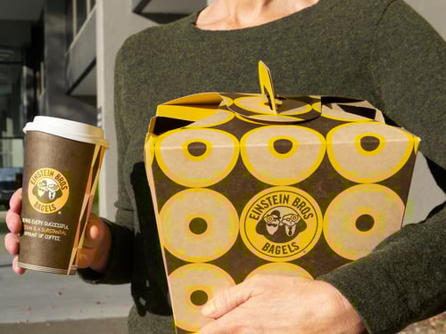 Einstein Bros. Bagels Model With A Good For Groups Bagel Box under their left arm coupled with a Large Hot Drip Coffee being held with the right hand.