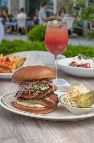 Fish sandwich plated at The Shed Restaurant sits on an outdoor patio table, featuring golden crispy fish on a toasted bun with fresh toppings, set against a relaxed open-air dining setting.